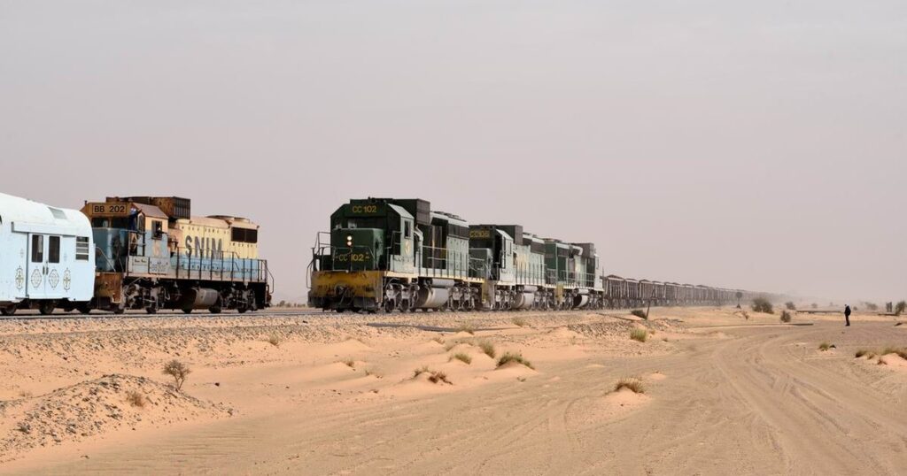 Un train de minerai de fer de la SNIM tiré par trois locomotives vertes traverse le désert mauritanien, à côté d'une locomotive bleue et jaune marquée SNIM